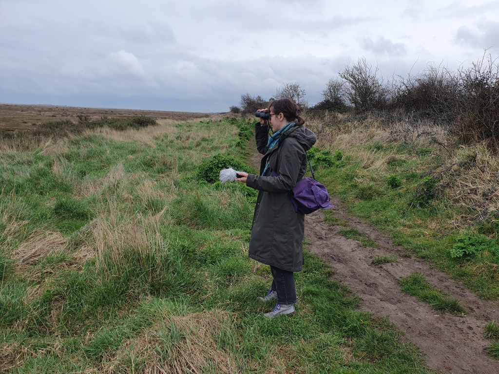 Flo is a white person wearing a green coat. They are next to a muddy path in a field, holding a recorder with a grey windjammer on it, and looking through a pair of binoculars.