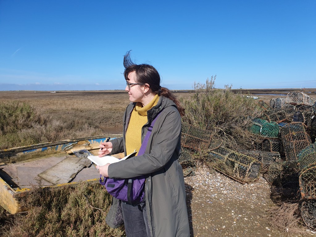 Flo is a white person wearing a yellow jumper and green coat. They are standing in front of a dinghy and crab pots on the Norfolk Coast while writing in a notebook.