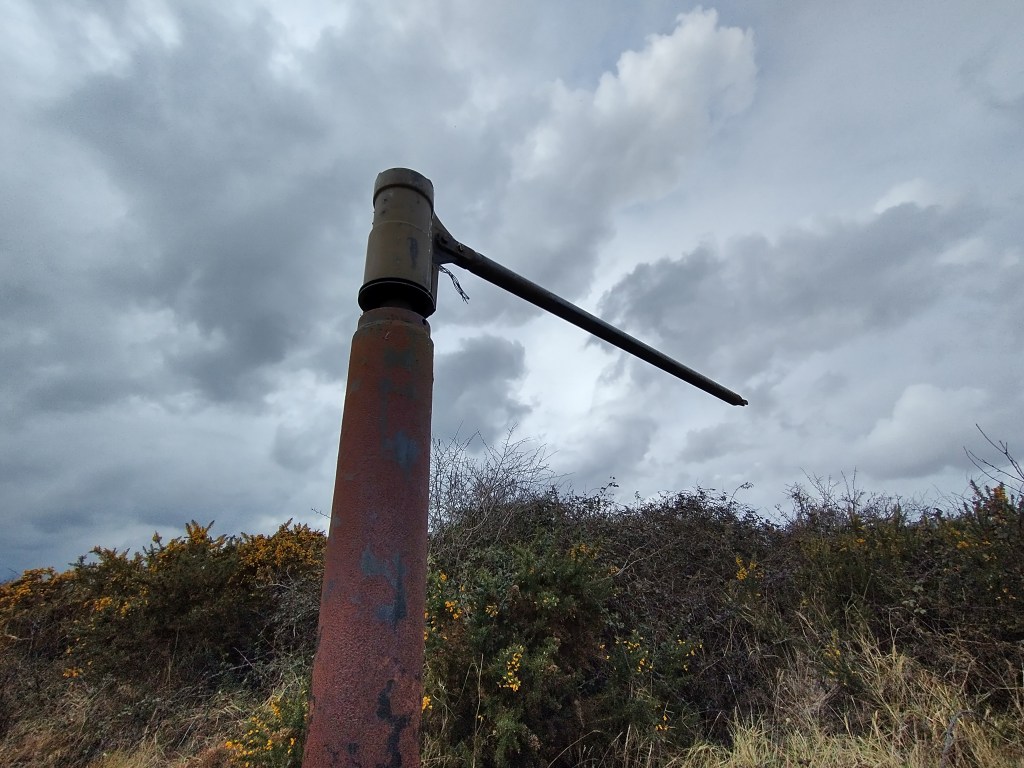 The Stiffkey Whirligig radio arm is a rusted metal pole with a smaller metal pole at its top forming a right angle. The radio arm is almost silhouetted against a grey cloudy sky and gorse bushes.