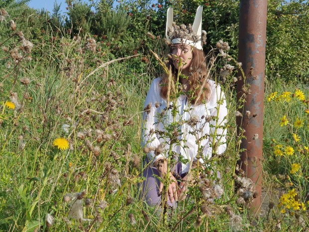 Flo is a white person with long brown hair, wearing a white shirt and a pair of handmade hare's ears on a headband. They are kneeling next to a rusted metal pole and are seen through the stems of summer wildflowers.