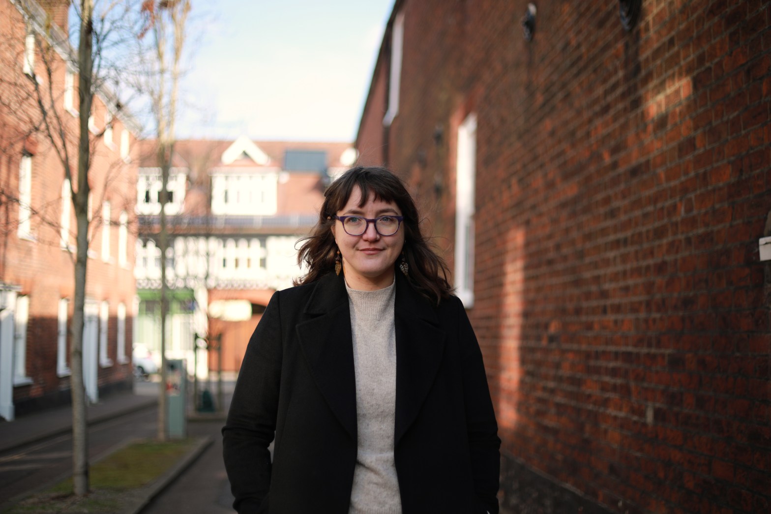 Flo is a white person with shoulder-length brown hair and a fringe. They are wearing purple-framed glasses, a cream jumper and a black coat and are standing in front of a blurred background of a narrow street with old red brick walls.